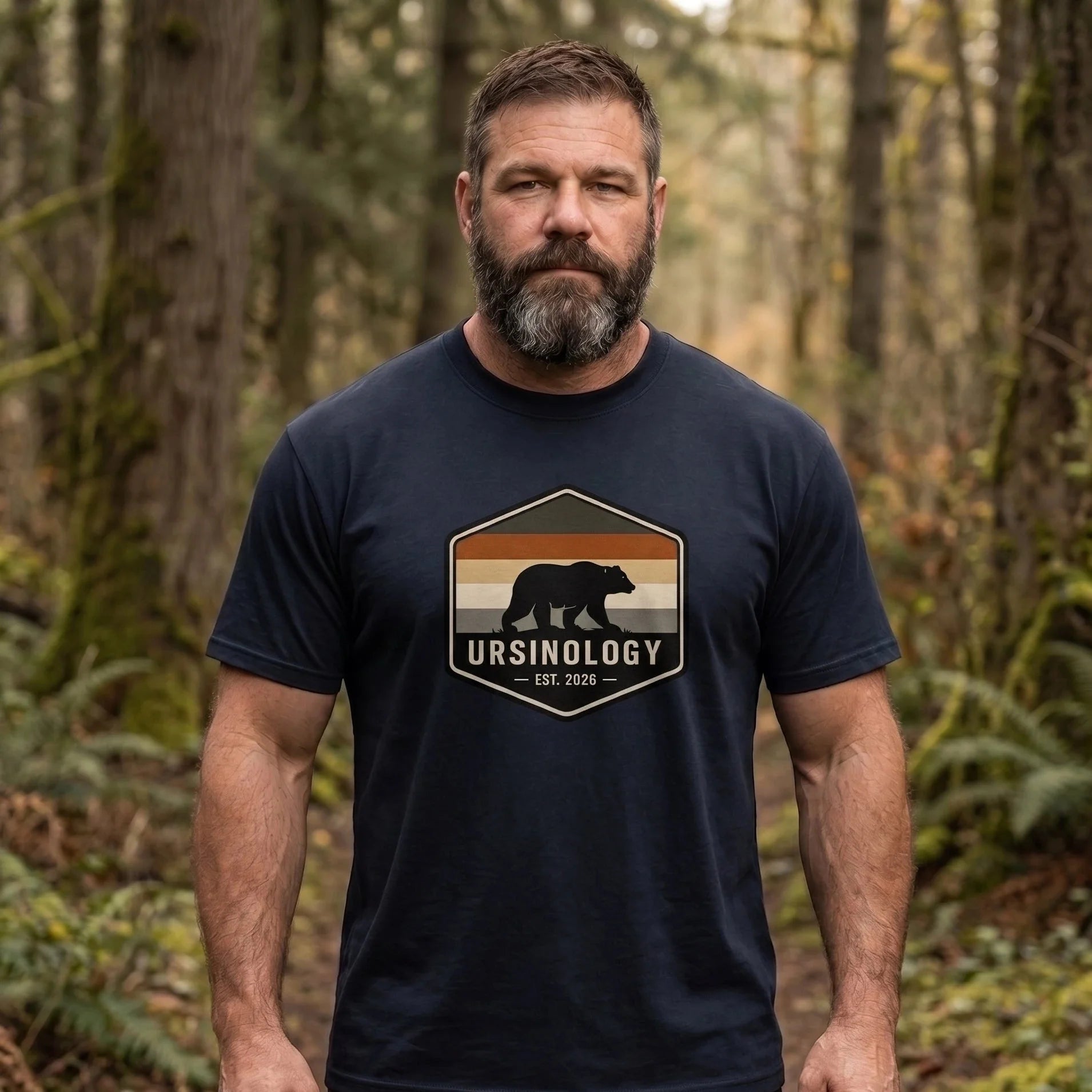 A bearded man wearing a navy blue t-shirt with a retro-style bear graphic and the word 'URSINOLOGY' printed on the chest, standing in a forest.